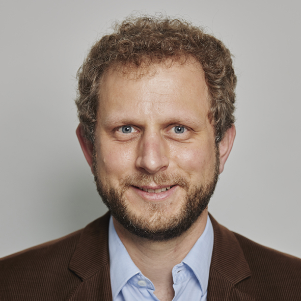 Portraitfoto von einem Mann mit lockigen dunkelblonden Haaren, blauen Augen und Bart vor einem grauen Hintergrund.