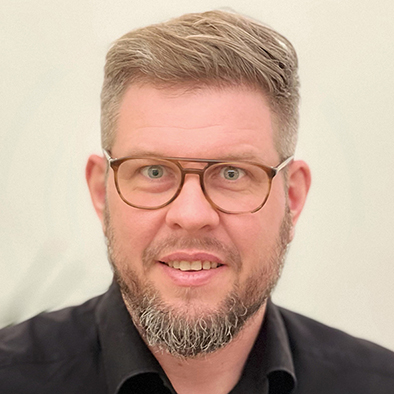 Ein Mann mit kurzen blonden Harren, Vollbart sowie brauner Brille ist in einer Portraitaufnahme zu sehen.