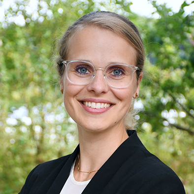 Eine Frau mit blonden Haaren, durchsichtiger Brille und einem schwarzen Blazer mit weißer Bluse drunter ist in einer Portraitaufnahme zu sehen.