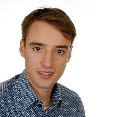 Ein Portraitfoto von einem Mann mit brauen Haaren und braunen Augen vor einer weißen Wand.