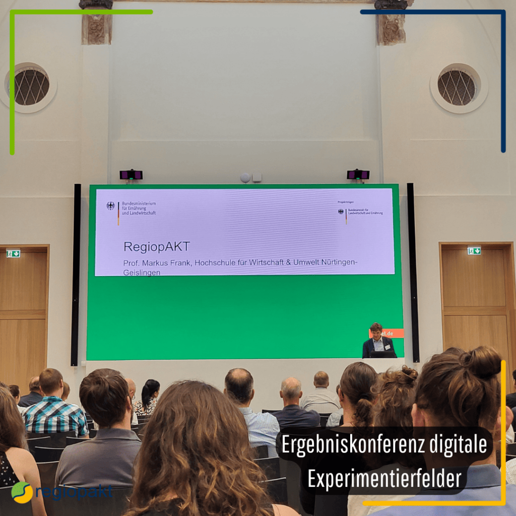 Große Leinwand auf der das Projekt regiopakt zu sehen ist. Vor der Leinwand steht ein Mann und davor sind Stuhlreihen mit sitzenden Menschen zu sehen.