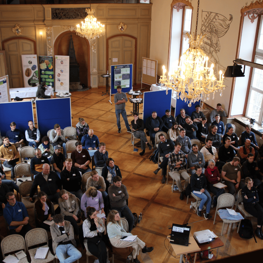 Zu sehen ist ein Saal aus einem Schloss, in dem in Stuhlreihen Menschen sitzen und zur Leinwand schauen. Im hinteren Bereich des Saals, sind verschiedene Pinnwände und Banner zu sehen.